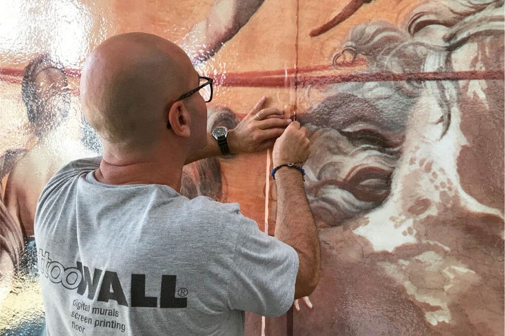 © Dekozone.com
Die dünne Membran des gedruckten Freskos wird wie eine Tapete aufgebracht. Mann bringt die dünne Membran wie eine Tapete auf die Wand auf.