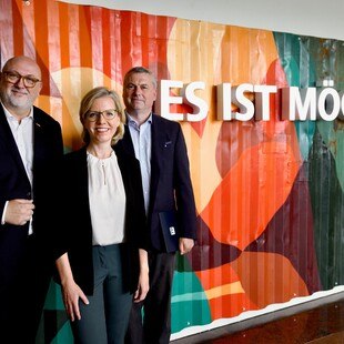 © ÖBB Scheiblecker  Günter Emberger, Leonore Gewessler und ein dritter stehen vor einer bunten Wand auf der steht: Es ist möglich